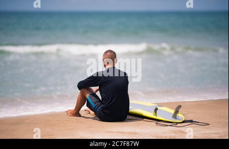 Junger Surfer mit Surfbrett, der alleine am Strand sitzt und auf das Meer blickt und auf die Wellen wartet. Stockfoto