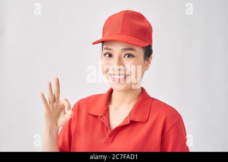 Portrait von attraktiven Lieferung Frau in roten T-Shirt und Mütze lächelnd und zeigt okey Zeichen isoliert auf rosa Hintergrund Stockfoto