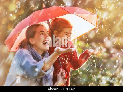 Fröhliche lustige Familie mit rotem Regenschirm unter der Herbstdusche. Mädchen und ihre Mutter genießen Niederschläge. Kinder und Mama spielen auf der Natur im Freien Stockfoto