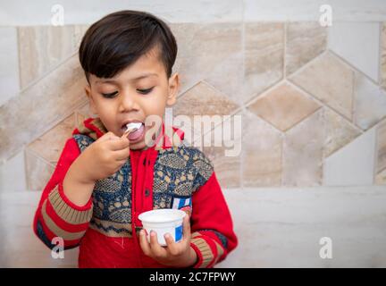 Niedlicher kleiner Junge in rotem Kleid, zu Hause Tasse Eis mit Löffel essen. Stockfoto