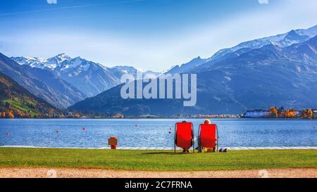 Spektakuläre Herbstansicht des Sees und Paar auf Stühlen sitzen in der Stadt Sell am See.. Fantastischer sonniger Tag über dem See. Ort: Zell am See, Salzburger Stockfoto