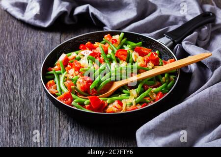 Grüne Bohnen mit Zwiebeln und Tomaten unter Rühren anbraten, in einer Pfanne auf einem Holztisch mit grauem Tuch frittieren, Landschaftsansicht von oben Stockfoto