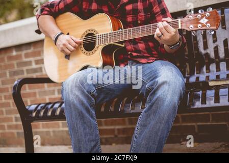 Männchen trägt einen roten und schwarzen Flanell, der auf einem sitzt Bank spielt Gitarre Stockfoto
