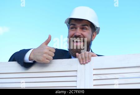 Mann mit Bart und breiten Lächeln trägt weißen Helm. Architekt in formellem Anzug auf blauem Himmel Hintergrund. Vorarbeiter schaut aus dem Holzzaun und zeigt Daumen nach oben. Bau- und Architekturkonzept Stockfoto