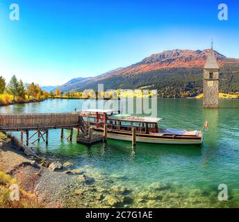 Fantastische Herbst Seenlandschaft mit einem Schiff in der Nähe der Pier und untergetauchten Glockenturm im See Reschen. Lage: Graun im Vinschgau Dorf, Reschensee Stockfoto