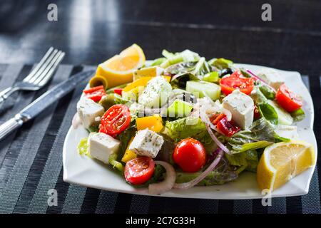 Gemüsesalat auf dem Teller, Tomaten, grüner Salat, Käse, Zitrone und Oliven, anderes Gemüse. Auf schwarzem Hintergrund. Stockfoto
