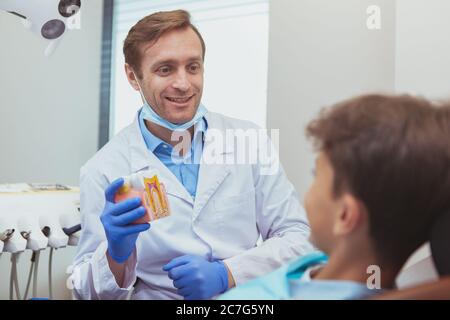 Reifen fröhlich männlichen Zahnarzt Ausbildung seiner jungen Patienten, zeigt Zahnmodell, erklärt Zahnpflege. Professioneller Zahnarzt im Gespräch mit einem kleinen Jungen Stockfoto