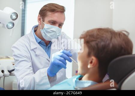 Professionelle Zahnmedizin, gesunde Zähne Konzept. Reifer Zahnarzt trägt medizinische Maske und Handschuhe, Untersuchung der Zähne eines kleinen Jungen Stockfoto