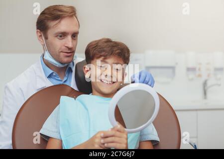 Charmanter, glücklicher kleiner Junge lächelnd, der in den Spiegel des Zahnarztbüros schaut. Reifer männlicher Zahnarzt und sein junger Patient überprüft Ergebnisse der zahnärztlichen Stockfoto