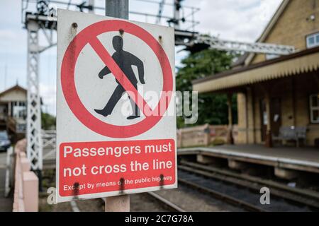 Nahaufnahme eines Metallwarnschildes für Passagiere, nicht um die Eisenbahnstrecke zu überqueren. Der Hintergrund zeigt einen verwagschwerten Bahnhof. Stockfoto