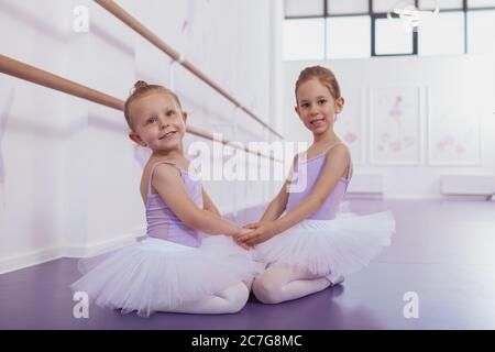 Schöne kleine Ballerinas halten Hände, sitzen auf dem Boden in der Ballettschule, lächeln zur Kamera. Glücklich liebenswert junge Ballett tanzen Mädchen restin Stockfoto