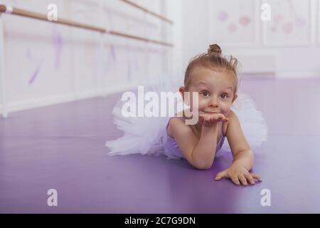 Charmante kleine Ballerina bläst Küsse in die Kamera, auf dem Boden liegend bei Ballettunterricht. Liebenswert kleines Mädchen im Ballett tanzen Outfit weht Kuss Stockfoto