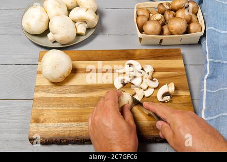 Mann Hände Cuttinde frische Champignon Pilze. Weißer und brauner Champignon auf grauem Holztisch in Scheiben geschnittene Champignons. Stockfoto