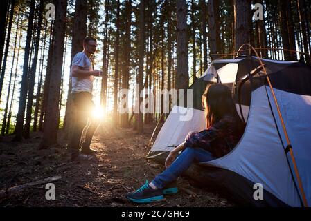 Abends Camping im Wald. Junge Frau, die in einem weißen Zelt sitzt und den Sonnenuntergang genießt. Auf Hintergrund Bäume und untergehende Sonne. Tourismus Abenteuer aktiven Lebensstil Konzept Stockfoto