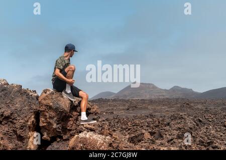 Junger Mann sitzt auf einem Lavastein, um den Timanfaya-Nationalpark zu beobachten. Lanzarote. Kanarische Inseln Stockfoto