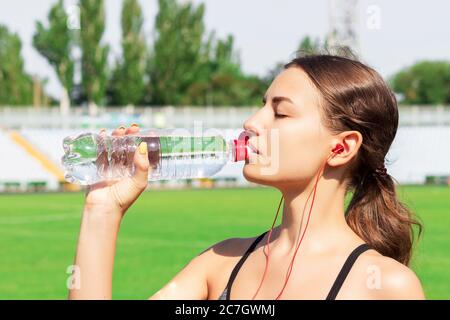 Schöne Frau trinkt Wasser und hört die Musik auf Kopfhörern im Stadion. Mädchen hat nach dem Training eine Pause. Stockfoto