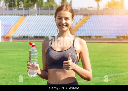 Schöne Frau trinkt Wasser und hört die Musik auf Kopfhörern im Stadion. Mädchen hat nach dem Training eine Pause. Stockfoto