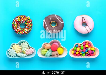Süßigkeiten - Lutscher, Baisenkerben, Makronen in Schale in Form von Wolke, Schokolade, mit bunten Belag Donut isoliert auf blauem Hintergrund Flat Lay Top View Stockfoto
