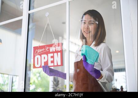 Asiatische Frau trägt Gesichtsschild Reinigung ihres Cafés vor dem Öffnen, während soziale Distanzierung und neue normale Stockfoto