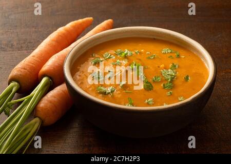 Karotten- und Koriandersuppe mit frisch gehacktem Koriander in einer braunen rustikalen Schüssel neben drei ganzen Karotten. Stockfoto