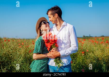 Liebevolles Paar zu Fuß in Mohn Feld mit Blumenstrauß. Glücklicher Mann und Frau umarmen lächelnd. Sommerurlaub Stockfoto