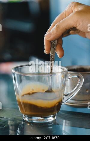 Nahaufnahme Hände einer Frau Rühren Kaffee, heiße Milch Kaffee tropft in Vietnam-Stil. Selektiver Fokus. Getränke- und Reisekonzept Stockfoto