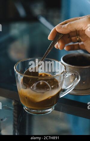 Nahaufnahme Hände einer Frau Rühren Kaffee, heiße Milch Kaffee tropft in Vietnam-Stil. Selektiver Fokus. Getränke- und Reisekonzept Stockfoto