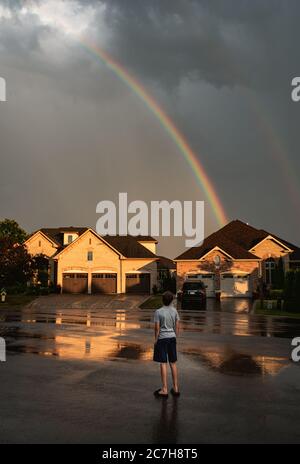 Junge, der auf der Straße steht und auf einen Regenbogen am Himmel blickt. Stockfoto