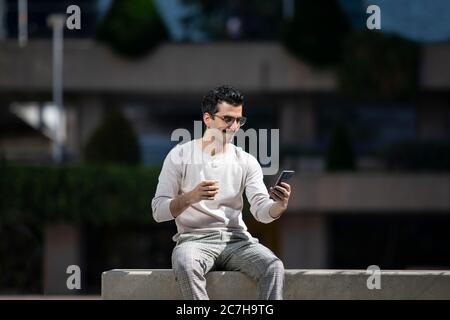 Junge Angestellte neben Wolkenkratzern, die Nachrichten auf dem Handy lesen und Kaffee trinken, während der Pause von der Arbeit Stockfoto