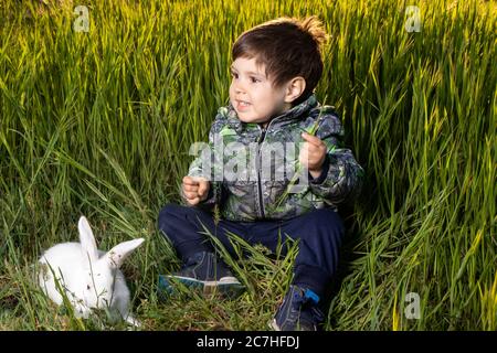 Kleiner Junge und Pannon White Hase sitzen auf grünem Gras. Freundschaft zwischen Mensch und Tier. Kinder und Haustiere. Stockfoto