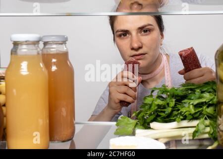 Das Mädchen schaut in den Kühlschrank und nimmt einen Stock geräucherter Wurst. Die Unterbrechung der Gewichtsverlust Ernährung. Stockfoto