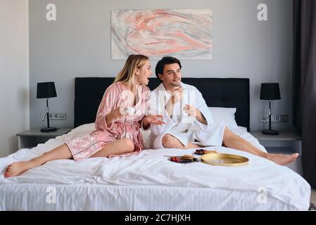Schöner Brünette Mann in weißem Bademantel und schöne blonde Frau in rosa velevt Robe glücklich mit Tassen Kaffee in den Händen reden. Junges Paar Stockfoto