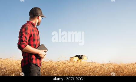 Millennial männlich Person mit Tablette sieht einen Erntemaschine in einem Weizen oder Roggen Feld. Moderner Landwirt, Landwirtschaft Business Management, lokale Unternehmer Stockfoto