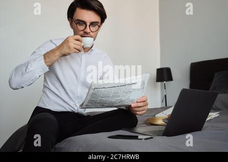 Junge schöne Brünette Mann in weißem Hemd und Brille verträumt Kaffee trinken und Zeitung lesen sitzen auf dem Bett mit Laptop und Handy in der Nähe Stockfoto