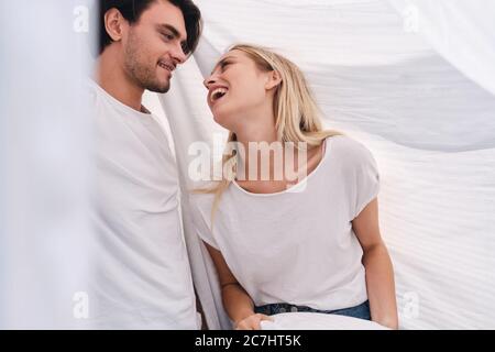 Junge lächelnde Brünette Mann und hübsche blonde Frau in weißen T-Shirts glücklich Blick auf einander unter Decke im Bett zu Hause sitzen Stockfoto