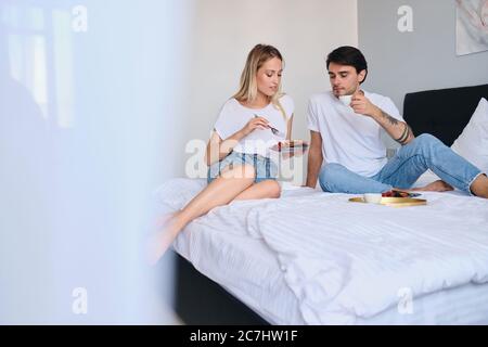 Nachdenklich Brünette Mann Kaffee trinken, während ziemlich blonde Frau in der Nähe essen Pfannkuchen mit Beeren. Junge attraktive Paar in weißen T-Shirts mit Stockfoto
