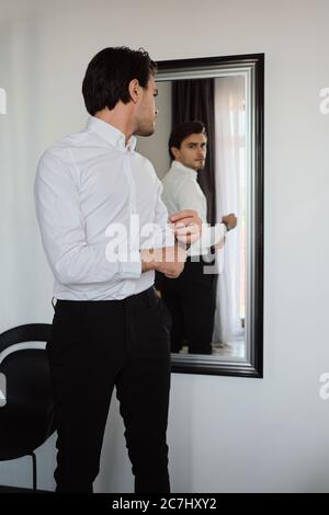 Junge schöne Brünette Mann in weißem Hemd und schwarzen Hosen traumhaft in Spiegel zu Hause suchen Stockfoto