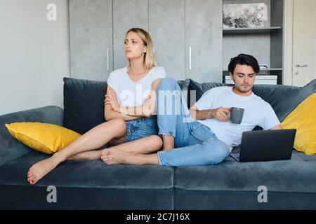 Junge beleidigte blonde Frau traurig beiseite schauen, während Brünette Mann mit Tasse Kaffee in der Hand nachdenklich arbeiten auf Laptop. Paar in schlechter Laune Stockfoto