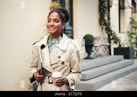 Schöne afroamerikanische Geschäftsfrau in stilvollen Trenchcoat mit Laptop und Kaffee zu gehen selbstbewusst die Straße hinunter Stockfoto