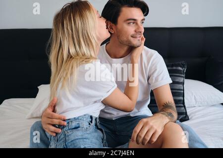 Attraktive Brünette Mann glücklich beiseite schauen, während ziemlich blonde Frau umarmt ihn. Junge schöne Paar in weißen T-Shirts sitzen auf dem Bett zusammen Stockfoto