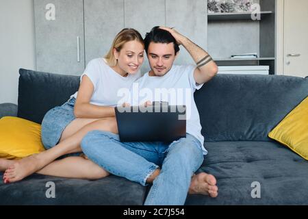 Ziemlich lächelnde blonde Frau und attraktive Brünette Mann glücklich mit Laptop zusammen. Junge schöne Paar sitzt auf dem Sofa in modernen Hause Stockfoto
