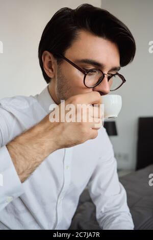 Nahaufnahme eines jungen attraktiven Brünetten-Mannes in weißem Hemd und Brille auf dem Bett sitzen und im Hotel traumhaft Kaffee trinken Stockfoto