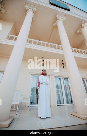 Schöner Mann aus dem Nahen Osten, arabischer muslim in traditioneller Kleidung, der vor dem weißen, schönen, modernen Haus steht Stockfoto