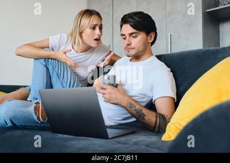 Traurige emotionale blonde Frau wütend im Gespräch mit ruhigen Brünette Mann mit Tasse Kaffee in den Händen und Laptop in der Nähe. Junges Paar in schlechter Stimmung sitzt auf dem Sofa Stockfoto