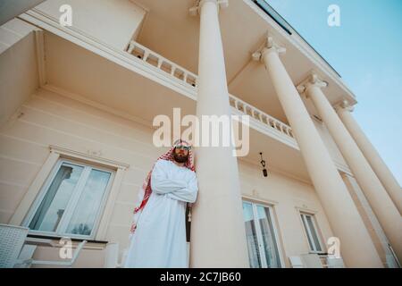 Schöner Mann aus dem Nahen Osten, arabischer muslim in traditioneller Kleidung, der vor dem weißen, schönen, modernen Haus steht Stockfoto