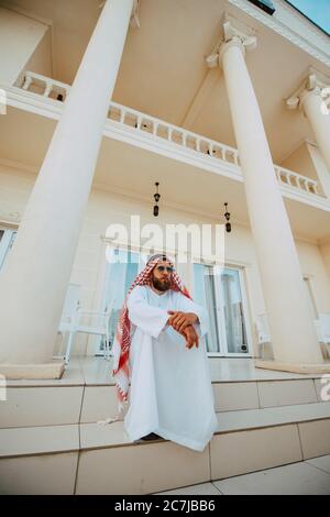 Schöner Mann aus dem Nahen Osten, arabischer muslim in traditioneller Kleidung, der vor dem weißen, schönen, modernen Haus steht Stockfoto