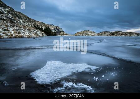 Das Meer ist mit Eis bedeckt, umgeben von Felsen und Häusern darunter Ein wolkig Himmel im Winter Stockfoto