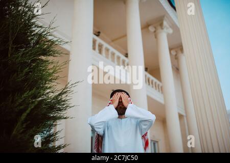 Schöner Mann aus dem Nahen Osten, arabischer muslim in traditioneller Kleidung, der vor dem weißen, schönen, modernen Haus mit den Händen über seinem Gesicht steht Stockfoto