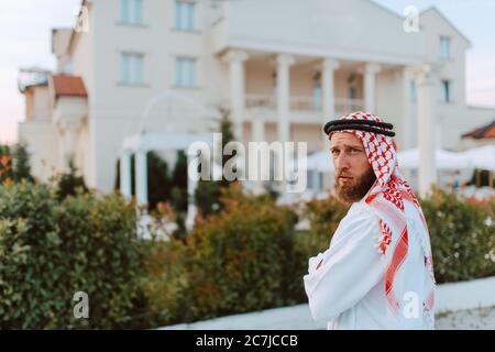 Schöner Mann aus dem Nahen Osten, arabischer muslim in traditioneller Kleidung, der vor dem weißen, schönen, modernen Haus steht Stockfoto