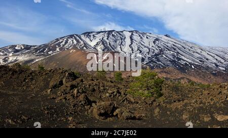 Ätna, Nationalpark Ätna, Parco dell –´Ätna, schneebedeckter Gipfel des Ätna im Hintergrund, roter Lavabugel in der Mitte, braune Lavasteine im Vordergrund, blauer Himmel, teilweise bewölkt Stockfoto